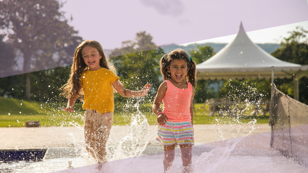 Two young girls laugh and run through a splash pad on a sunny summer day, surrounded by water sprays, illustrating safe and joyful outdoor summer play for children.